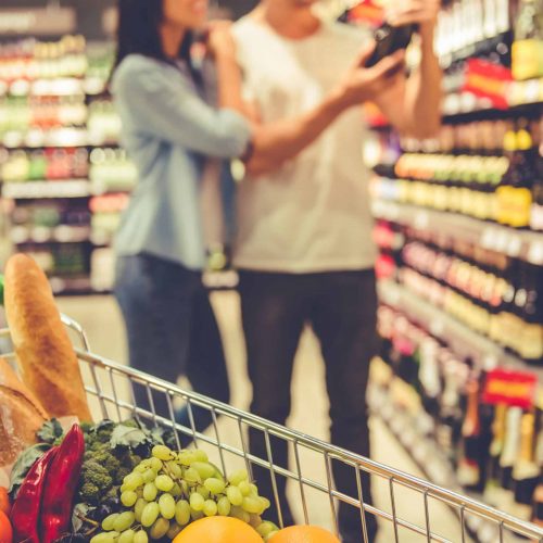 A shopping cart full of food, in the background beautiful young couple is choosing a beverage in the supermarket