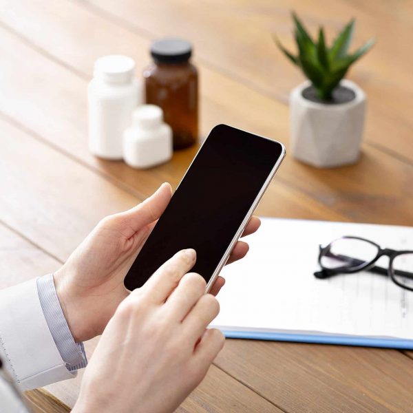 Mobile app and support patients online remotely. Lady middle aged doctor typing on smartphone with blank screen at workplace on wooden table with medicine jars with notepad and glasses, cropped
